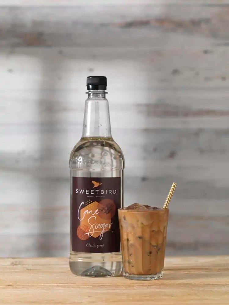 Bottle of Sweetbird Cane Syrup with a glass of iced coffee on a wooden surface.