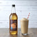 Bottle of Sweetbird English Toffee syrup next to a glass of a creamy drink on a wooden surface.